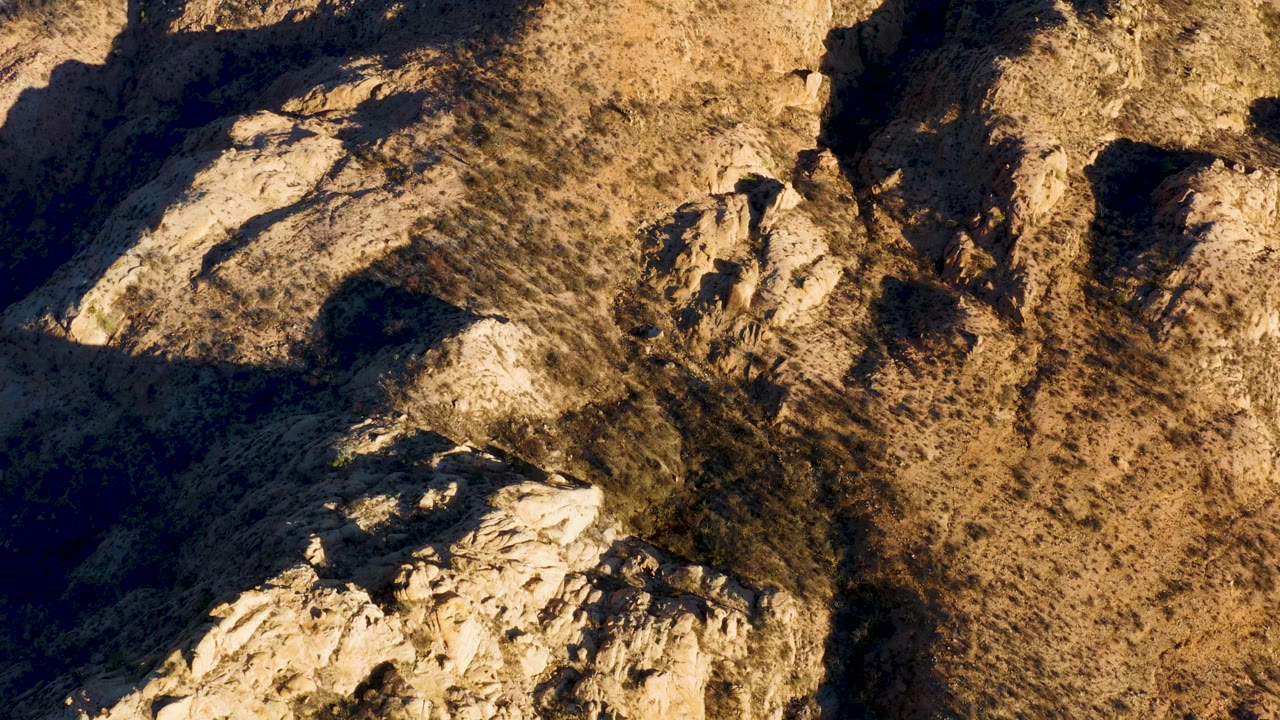 Aerial frame over Cabo Pulmo dry mountain ridge, with pale rock formations at lower left, scrub-dotted tan ground, heavy side shadow, and no visible people.