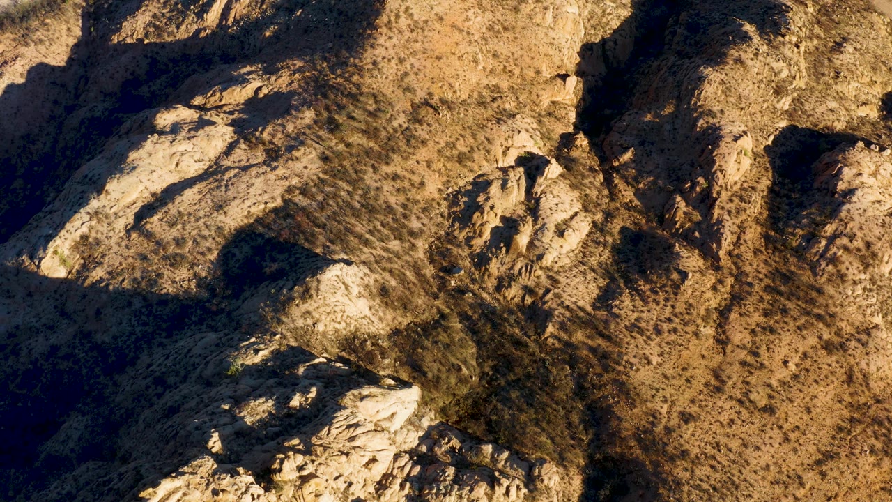 Mid-clip Cabo Pulmo terrain view with jagged pale outcrops, dry brush, brown hillside, dark recessed pockets, and no visible people.