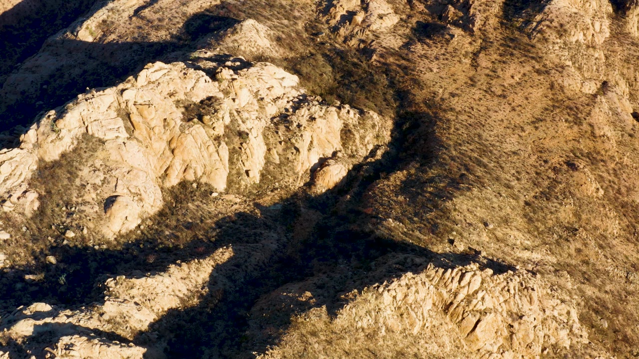 A Cabo Pulmo aerial frame with a pale rocky ridge near the center-left, dry brown hillside texture to the right, and deep shadows along the lower edge, no visible people.
