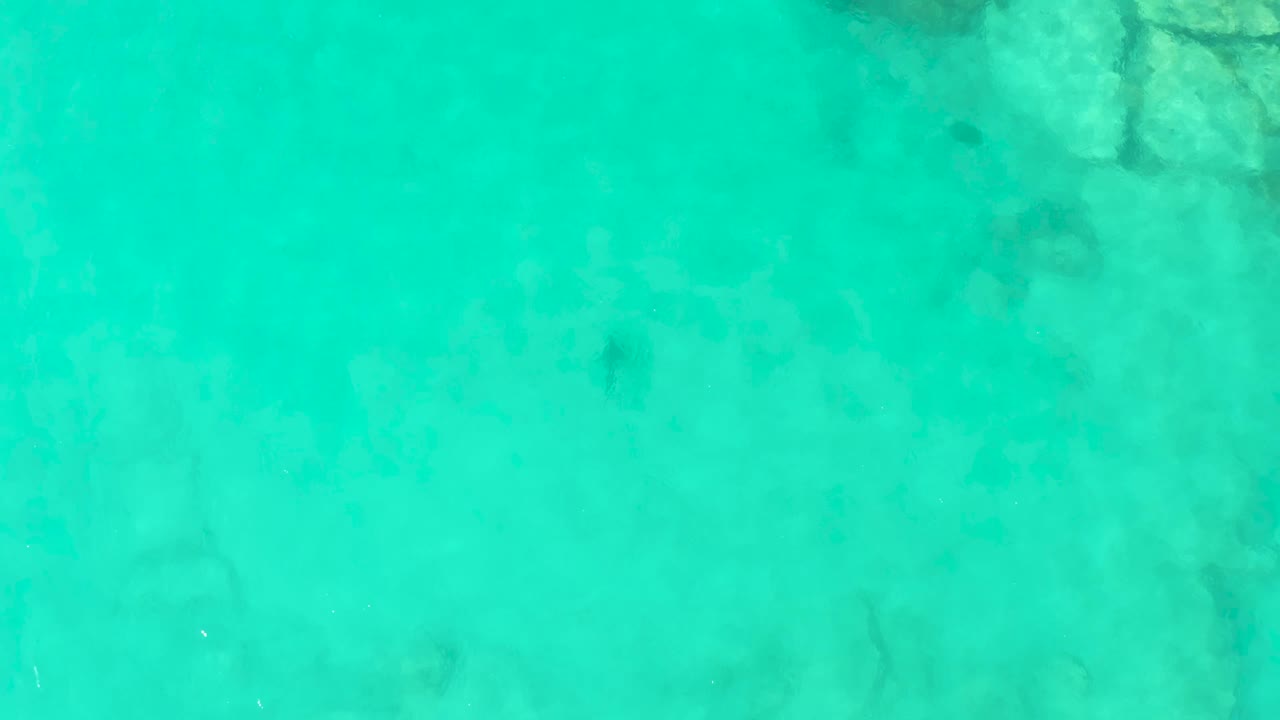 Shallow turquoise sea covers a rocky seabed in Cabo Pulmo, Baja California Sur, with darker patches visible through the water and no visible people.