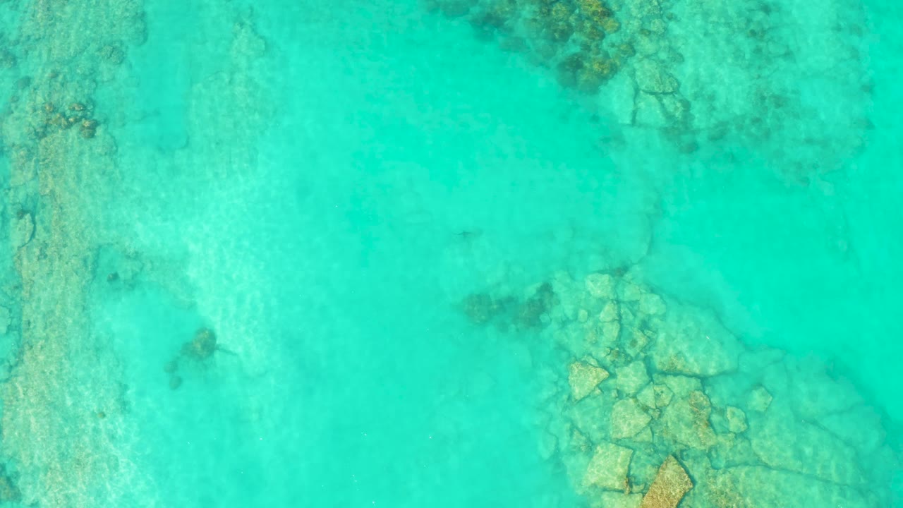 Overhead view in Cabo Pulmo of clear turquoise water revealing pale submerged rocks near the lower right, with no visible people.