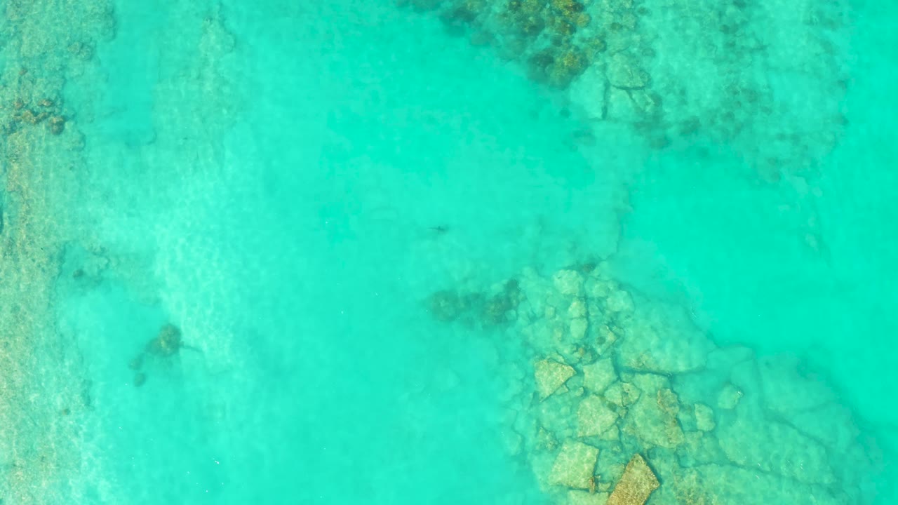 Bright turquoise water in Cabo Pulmo reveals a larger submerged rock formation in the lower right and faint reef texture near the top, with no visible people.