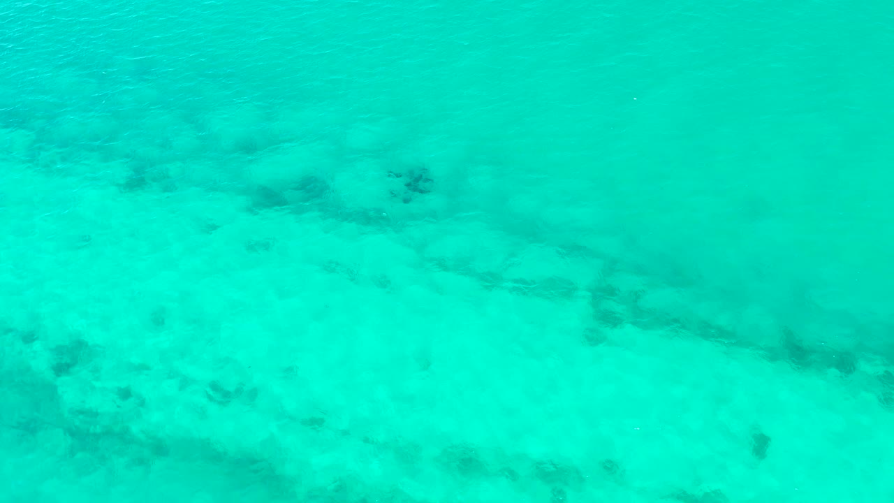 Turquoise water fills the frame above the Cabo Pulmo seabed, where pale sand and uneven dark patches remain visible through the rippled surface with no visible people.