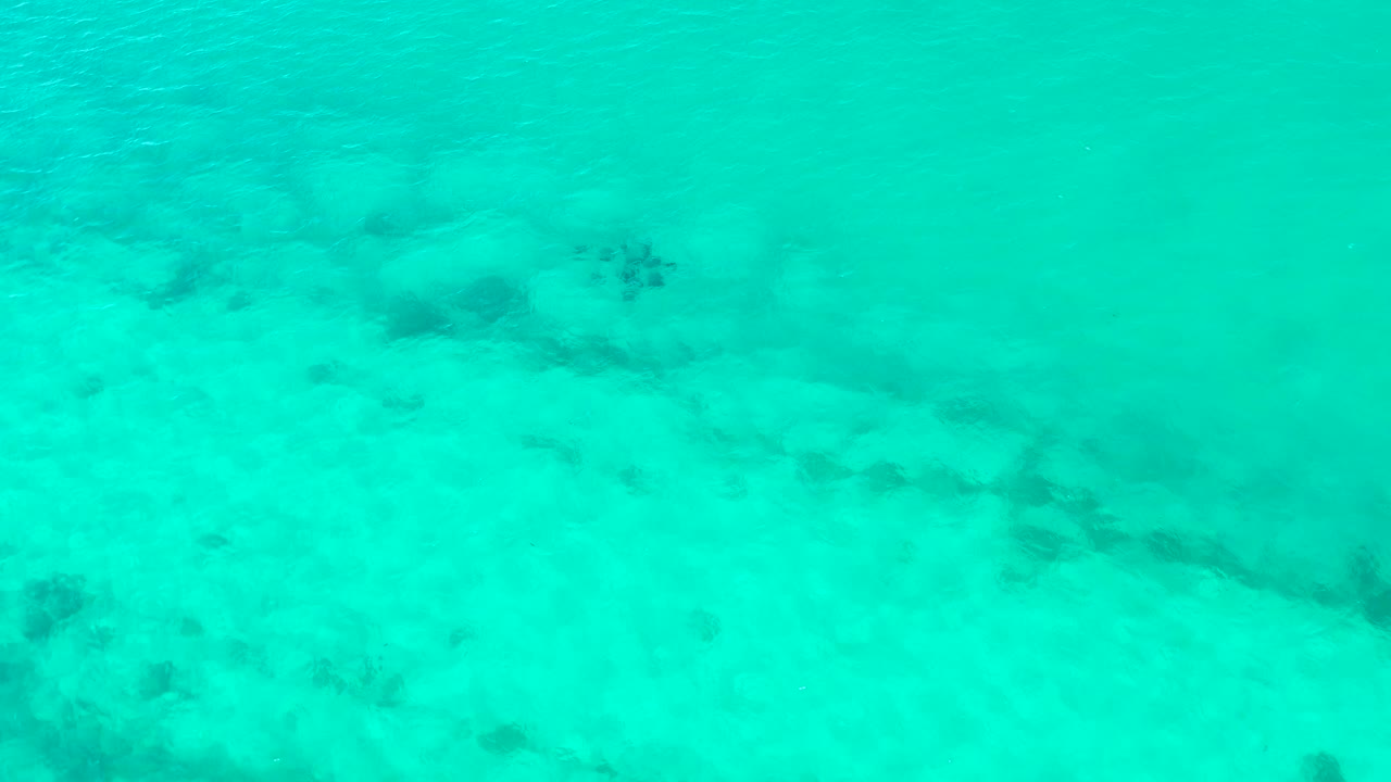 Clear turquoise water over Cabo Pulmo shows the sandy ocean floor beneath, with darker coral or rock patches spread across the lower and upper areas and no visible people.