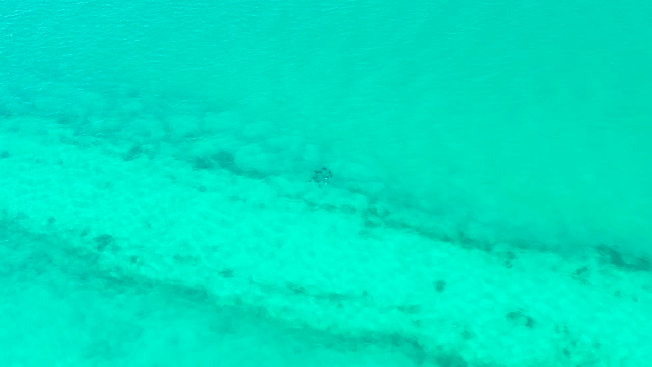 Clear blue-green water covers the sandy Cabo Pulmo seabed, with a darker diagonal patch visible beneath the calm surface and no visible people.