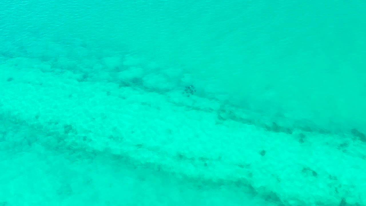 A shallow turquoise ocean-floor view in Cabo Pulmo shows mottled sand and small dark areas beneath lightly rippled water, with no visible people.