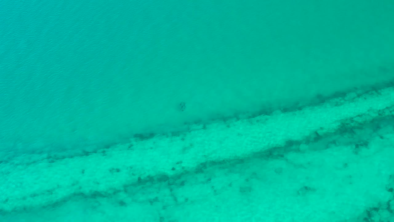 Overhead Cabo Pulmo water view one second in, where soft ripples pass over a broad pale seabed band and darker underwater markings, with no visible people.