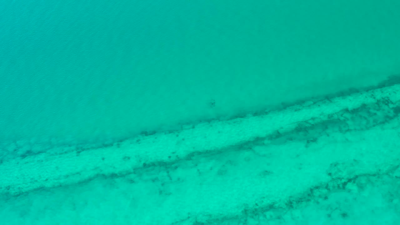 Mid-clip overhead view of blue-green Cabo Pulmo water shows the sandy bottom and scattered dark coral-like patches under a soft surface haze, no visible people.