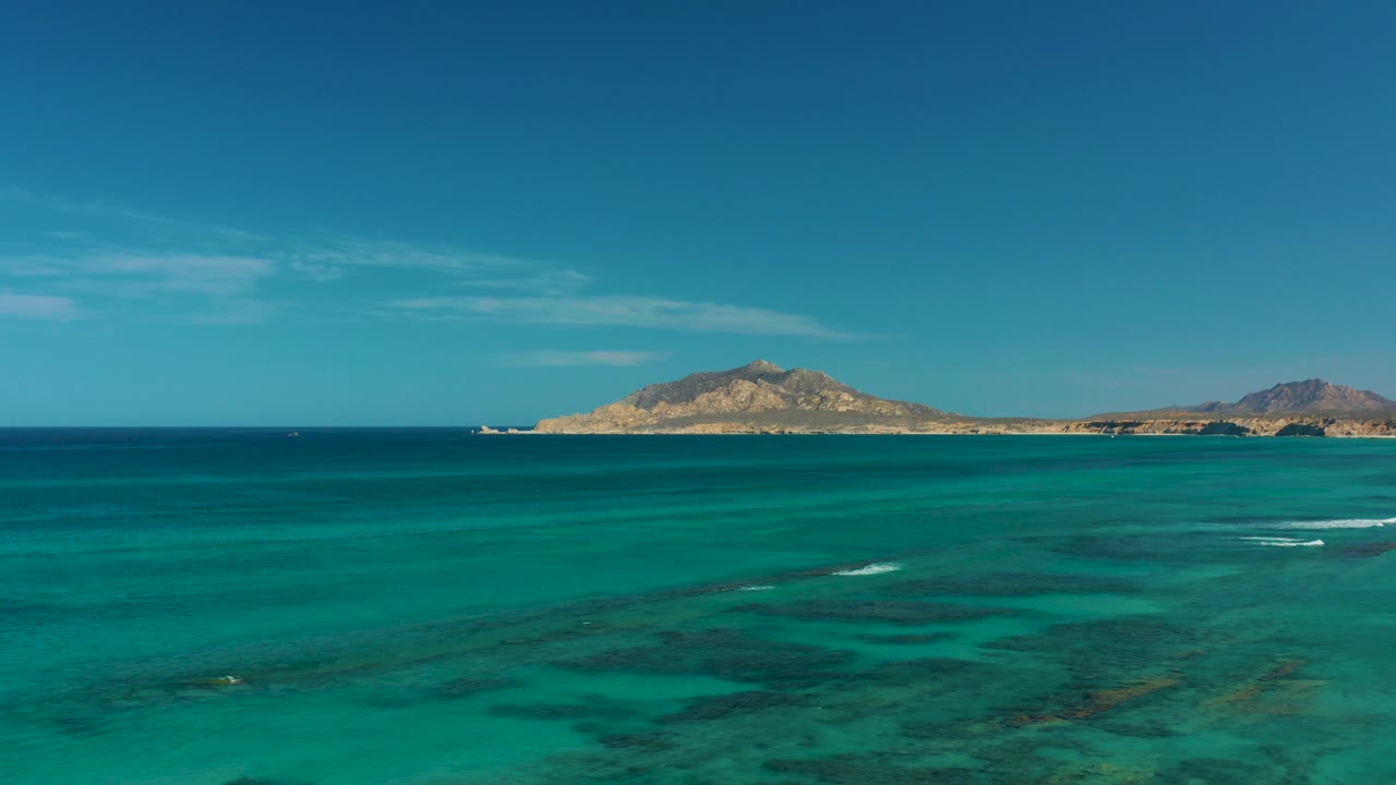 Wide aerial view of turquoise water near Cabo Pulmo, Mexico, with dark seabed patches, distant island hills, and no visible people.