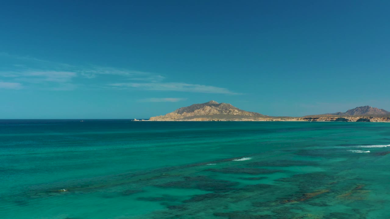 Aerial coastal frame near Cabo Pulmo showing transparent blue-green water, scattered whitecaps, low island terrain, and no visible people.