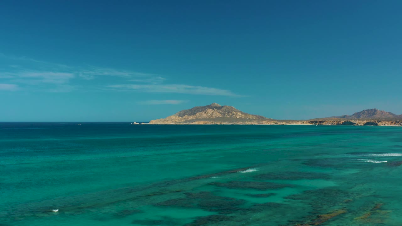 Wide drone-like view over shallow turquoise sea at Cabo Pulmo, with darker underwater areas, small breaking waves, distant hills, and no visible people.