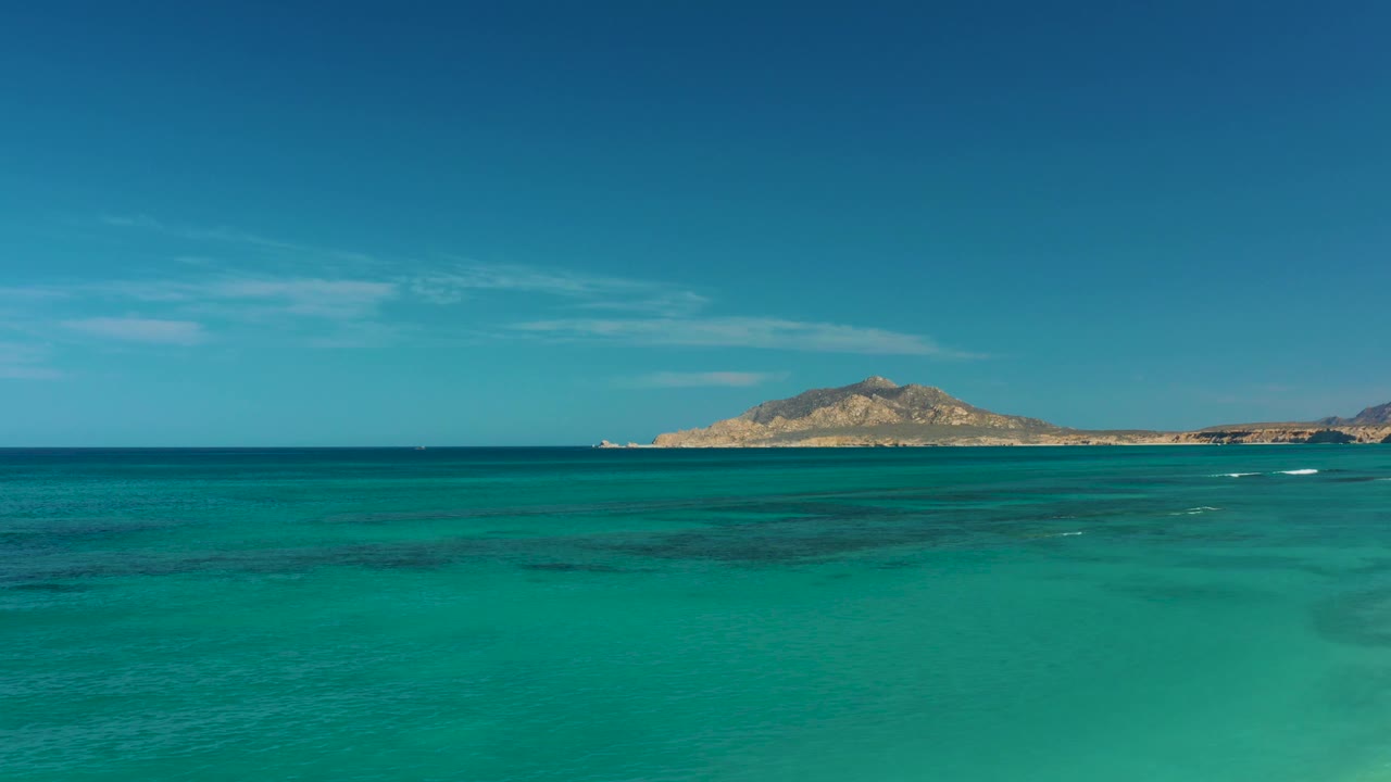 Clear turquoise water fills the foreground in Cabo Pulmo while faint white wave lines appear below the distant island, with no visible people.