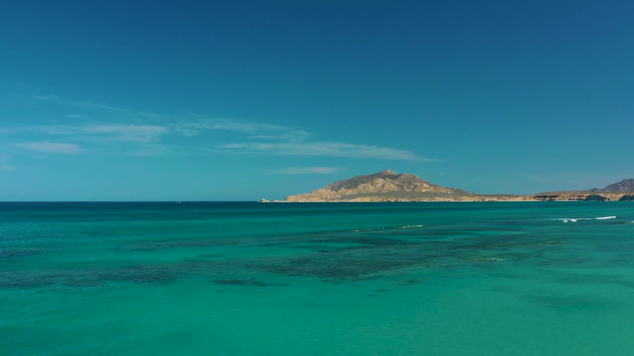 Turquoise coastal water in Cabo Pulmo shows several thin white foam streaks below the horizon, with the island centered in the distance and no visible people.