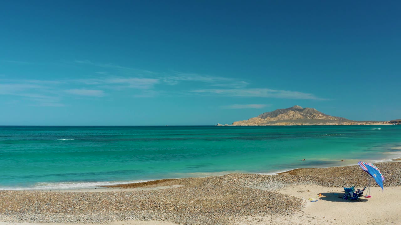 Cabo Pulmo beach frame with the umbrella and beach gear near the right edge, a curving pale shoreline, clear turquoise water, and low land across the horizon.