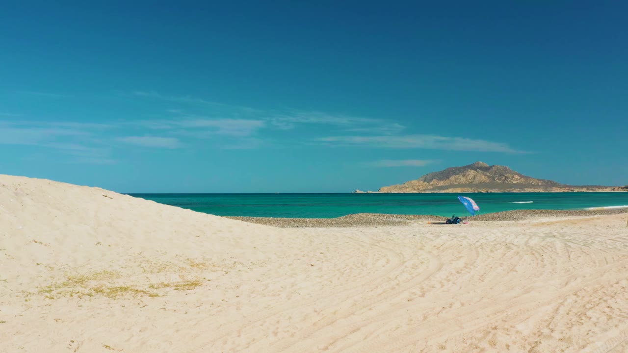 Cabo Pulmo beach view from a sandy dune, with turquoise water, a distant rocky landform, a blue umbrella near the shoreline, and no visible people.