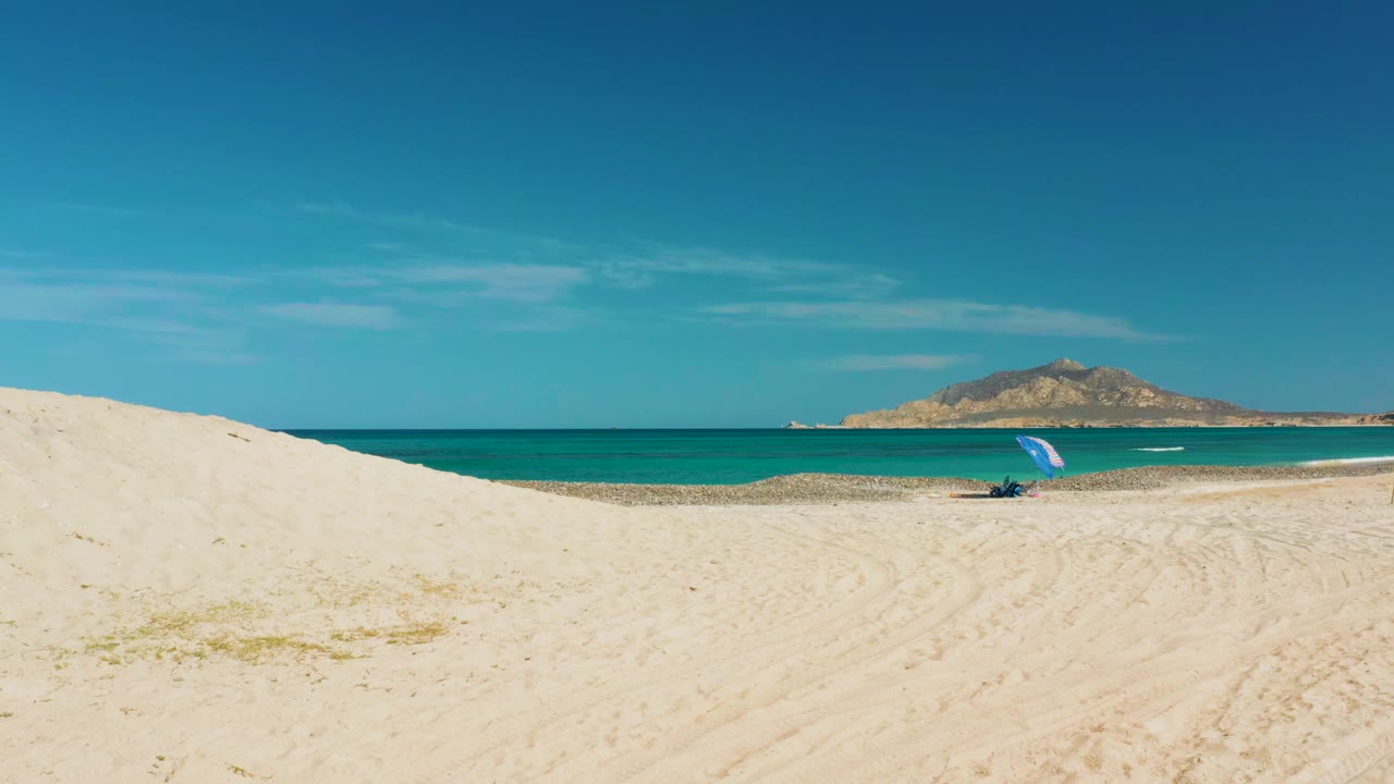 Cabo Pulmo sand dune ridge fills the lower frame while the turquoise ocean, rocky landform, and small blue umbrella remain visible, with no visible people.