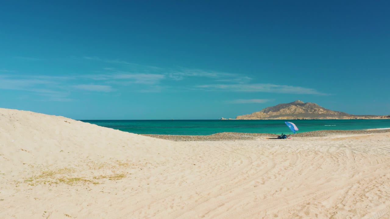 The Cabo Pulmo shoreline stays centered behind pale sand, with the blue umbrella closer to the right side of frame and no visible people on the beach.