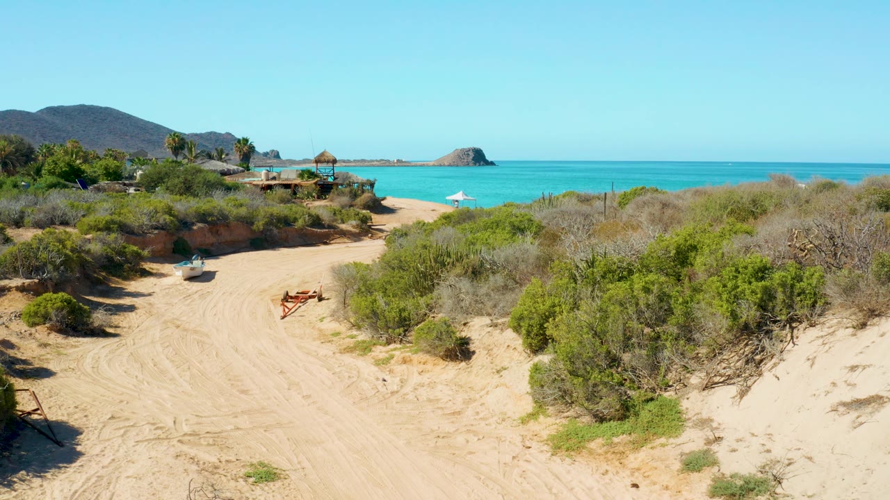 A wider section of the Cabo Pulmo sandy track passes through scrubby dunes toward the beach, with the ocean horizon, distant hill, and no visible people.