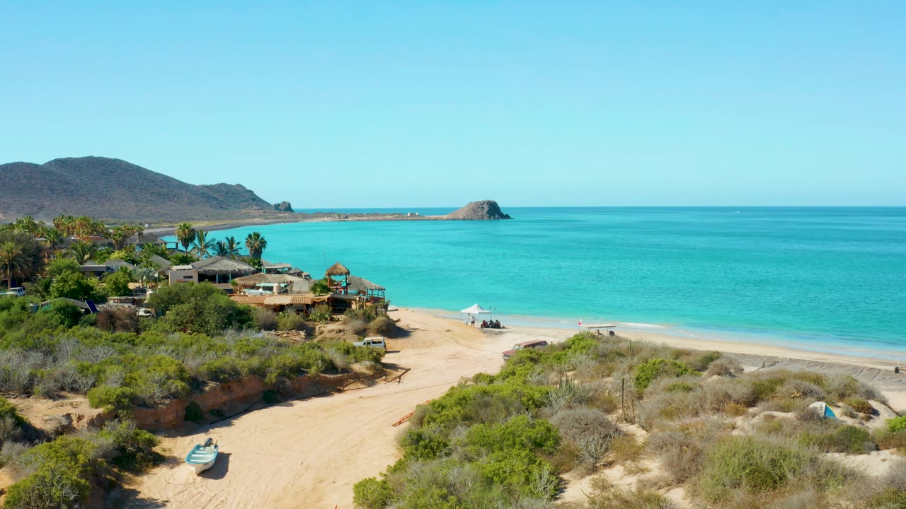 An elevated view in Cabo Pulmo shows a sandy track through dry scrub toward turquoise water, low buildings, distant mountains, and no visible people.