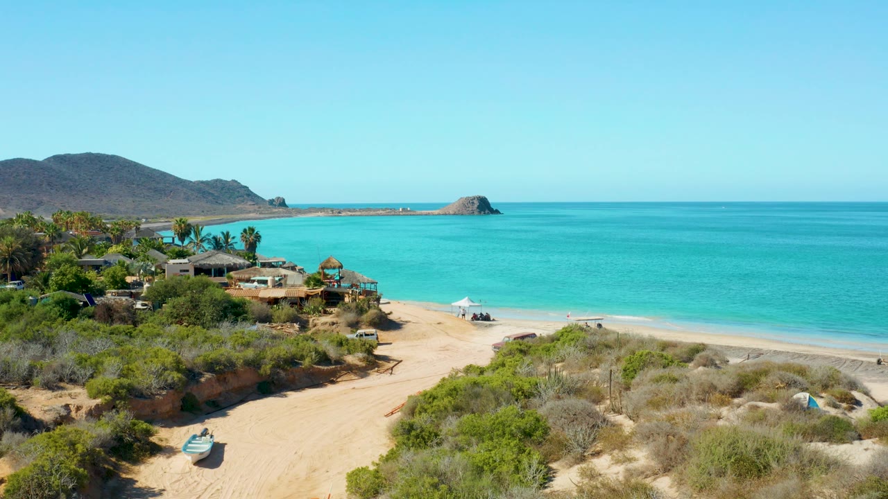 In Cabo Pulmo, the sandy access path and scrub dunes fill the foreground while turquoise beach water, low buildings, distant hills, and no visible people sit beyond.
