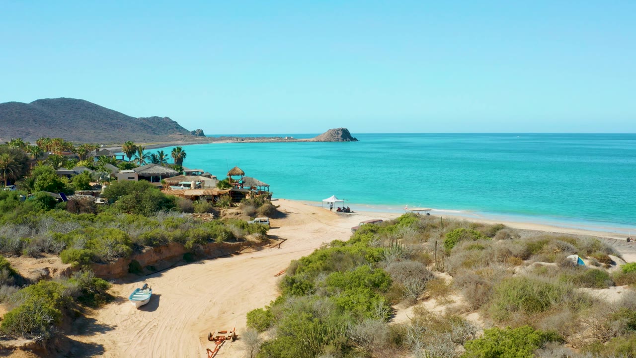 The Cabo Pulmo shoreline appears beyond a sandy path and dry vegetation, with turquoise water, a small offshore rock or island, low buildings, and no visible people.