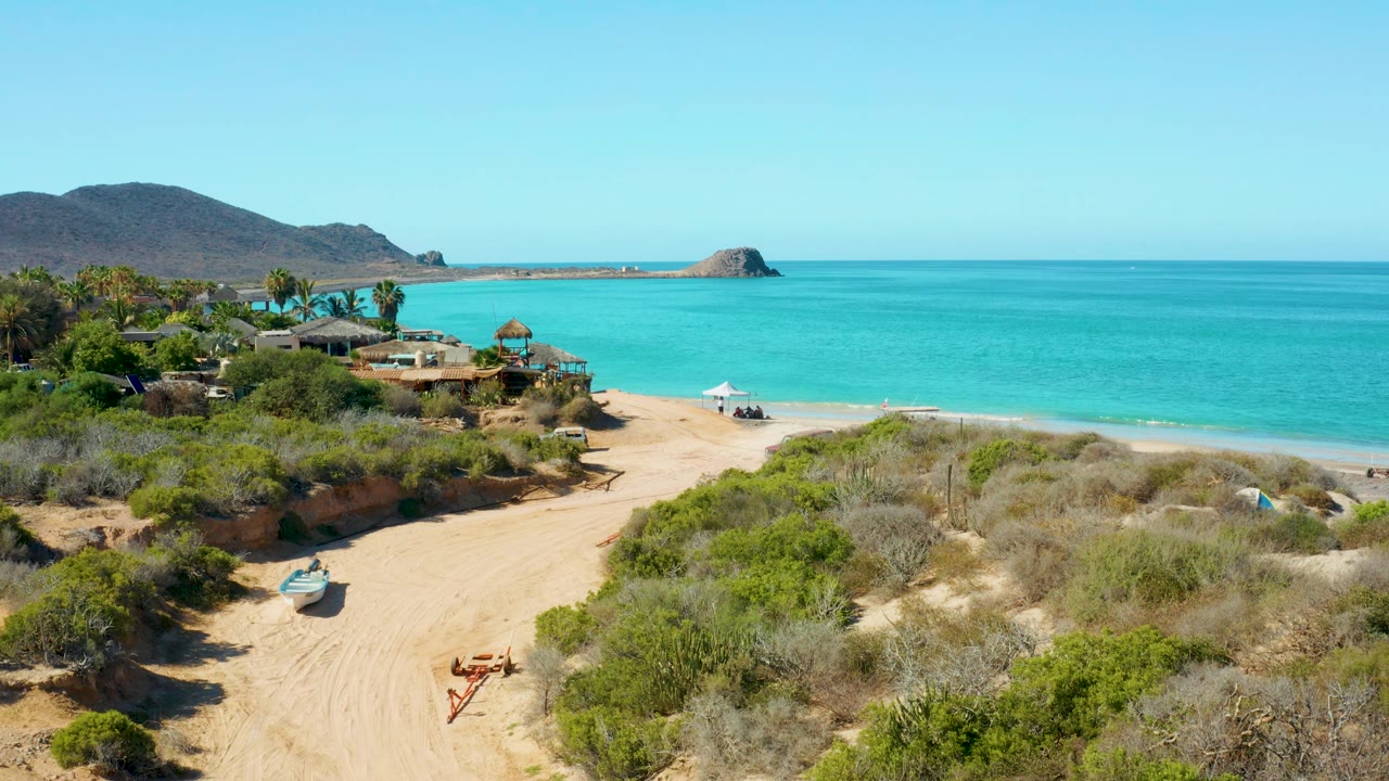 Wide view of sandy beach track, scrub, ocean, buildings, and mountains.