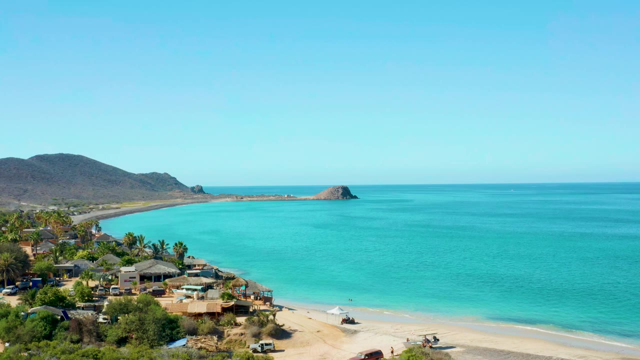 Cabo Pulmo coastal aerial view with turquoise water, pale beach, shoreline buildings, dry hills, and a rocky islet offshore, with no visible people.