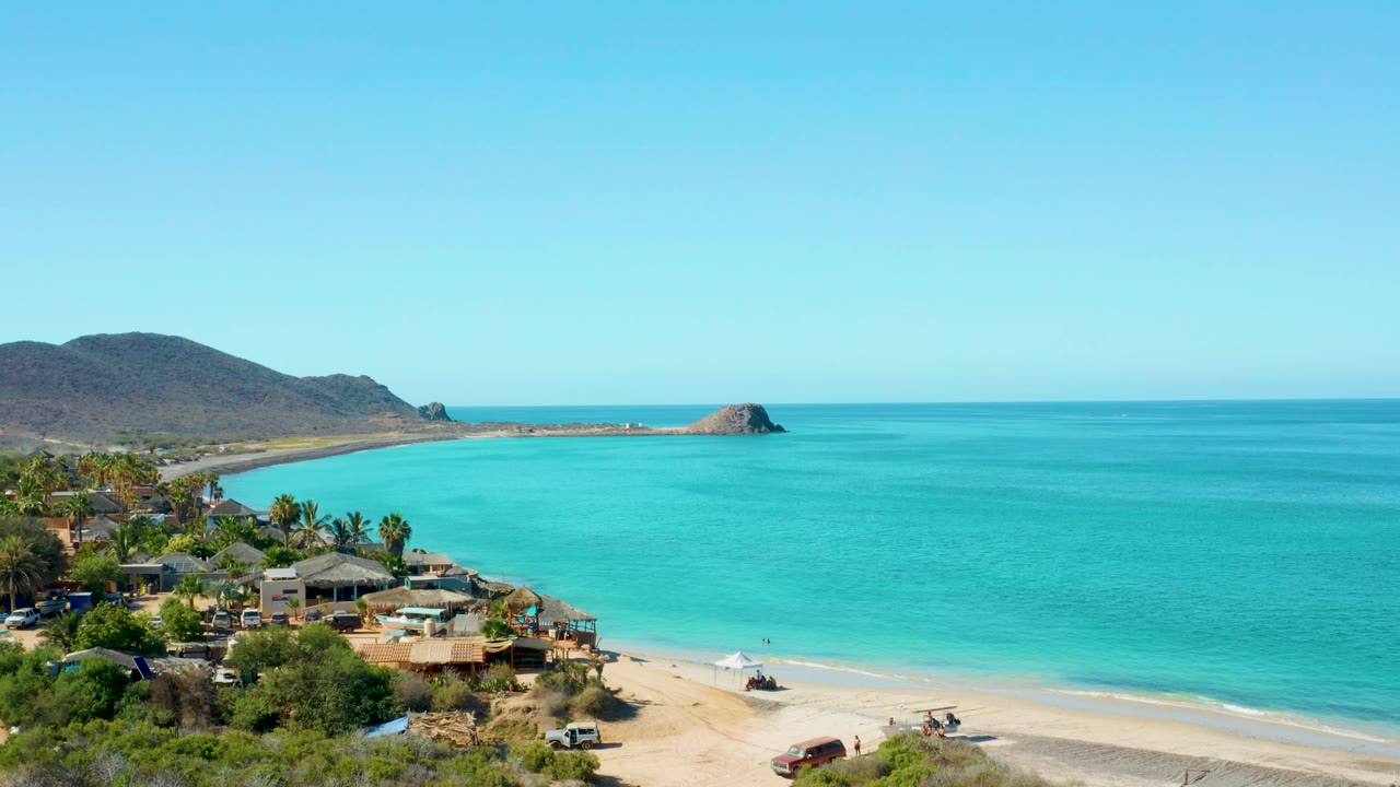 Cabo Pulmo aerial frame showing the beach curve, clustered shoreline buildings, turquoise bay, hazy horizon, and rocky islet near the hill-backed coast, with no visible people.