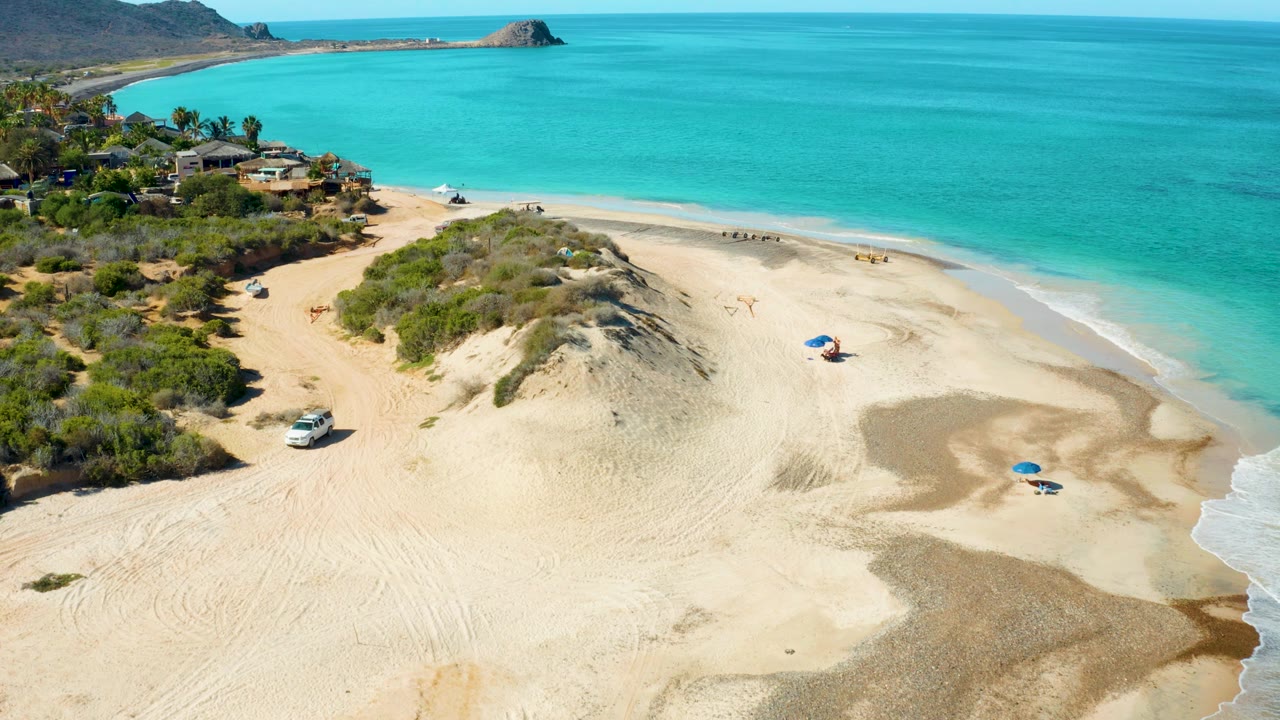 Wide Cabo Pulmo drone view with the sandy dune ridge filling the center, the white vehicle near the left sand track, small people near umbrellas, and bright turquoise ocean beyond the beach.
