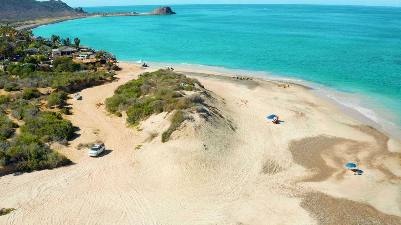 Drone view over beach dunes with a white vehicle and ocean shoreline.