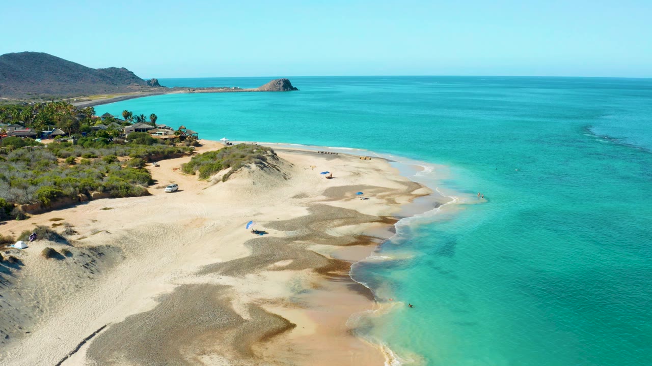 Wide aerial view over Cabo Pulmo beach with pale dunes in front, turquoise ocean to the right, vegetation and homes to the left, and low hills along the horizon.