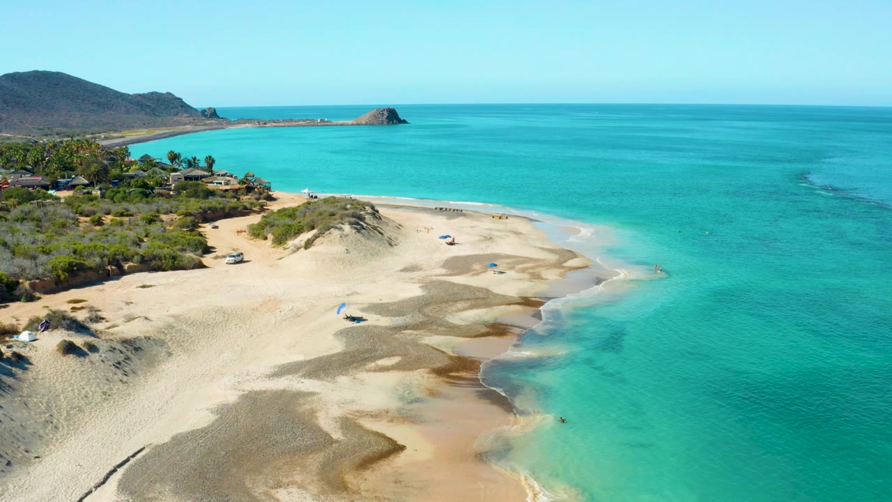 Drone view of beach dunes beside clear turquoise water and small umbrellas.