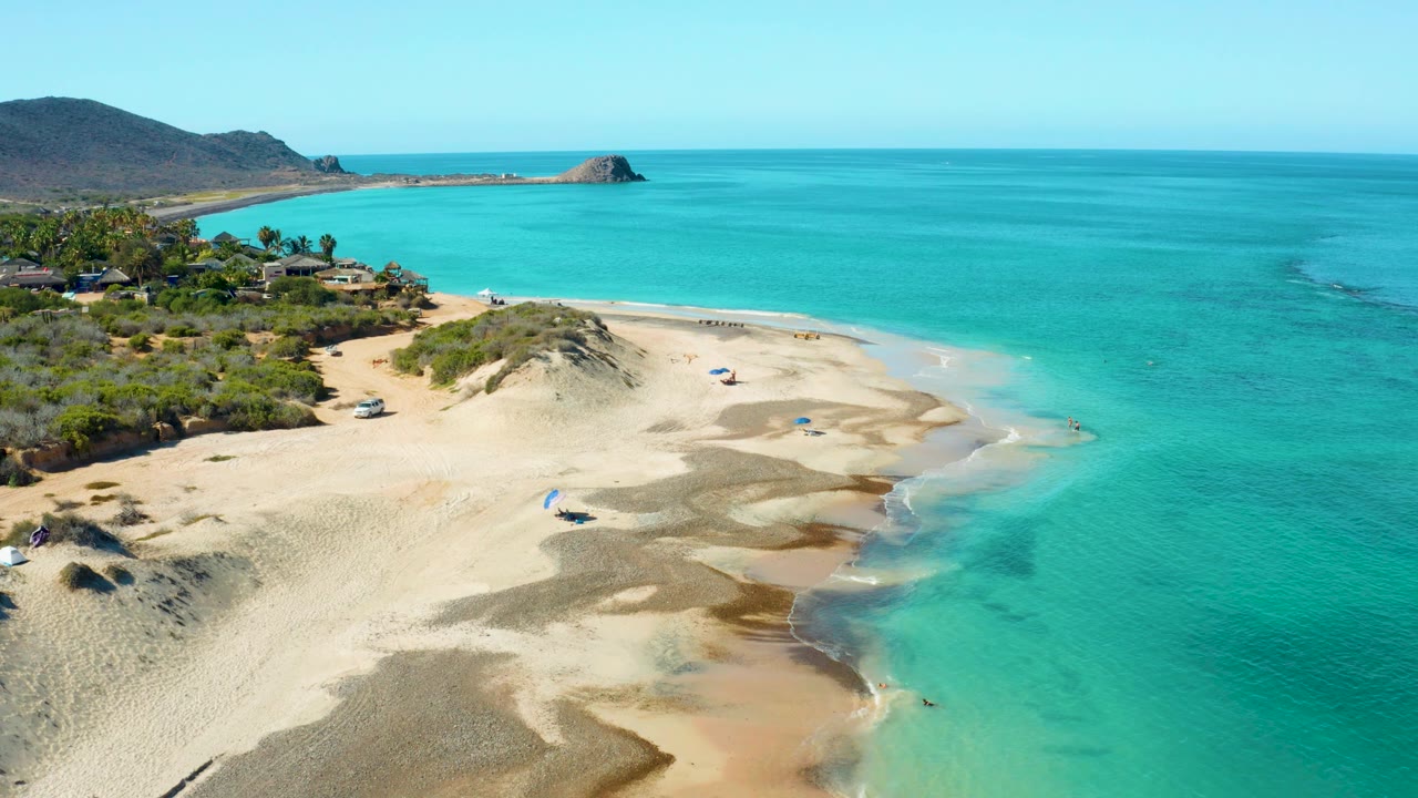 Cabo Pulmo aerial view with the sandy spit and surf line more prominent, a few tiny beachgoers near the water, umbrellas on the sand, and low hills behind the bay.