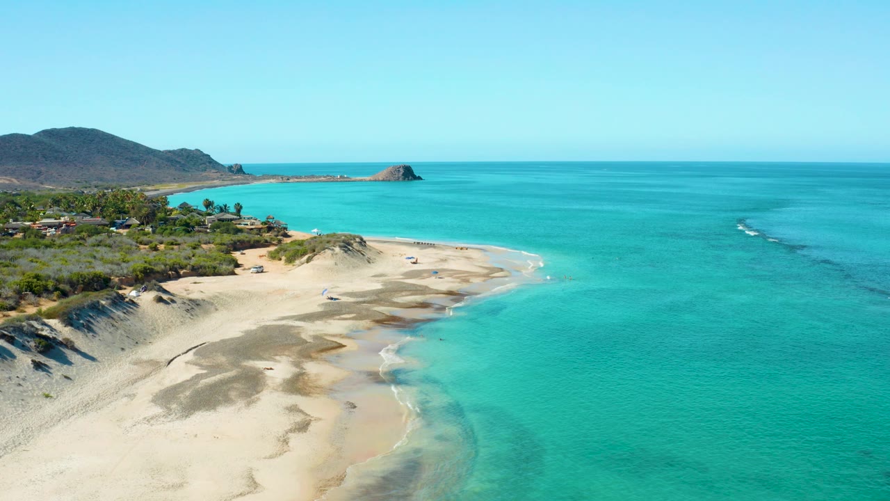 Cabo Pulmo shoreline from a high angle, with sand ridges filling the lower frame, clear water along the right edge, low vegetation at left, and no visible people.