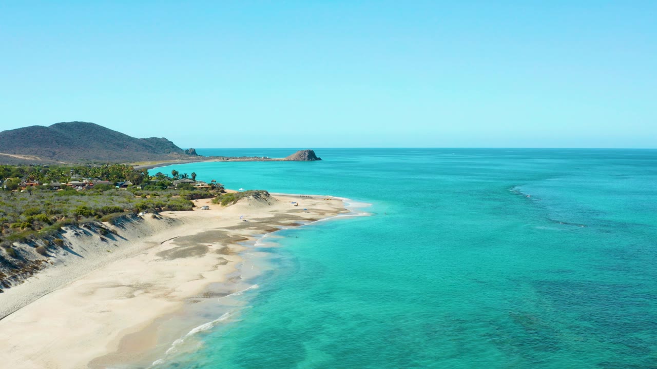 Aerial view over Cabo Pulmo showing a pale sandy beach curving into clear turquoise water, with low hills, greenery, scattered buildings, and no visible people at the start of the clip.