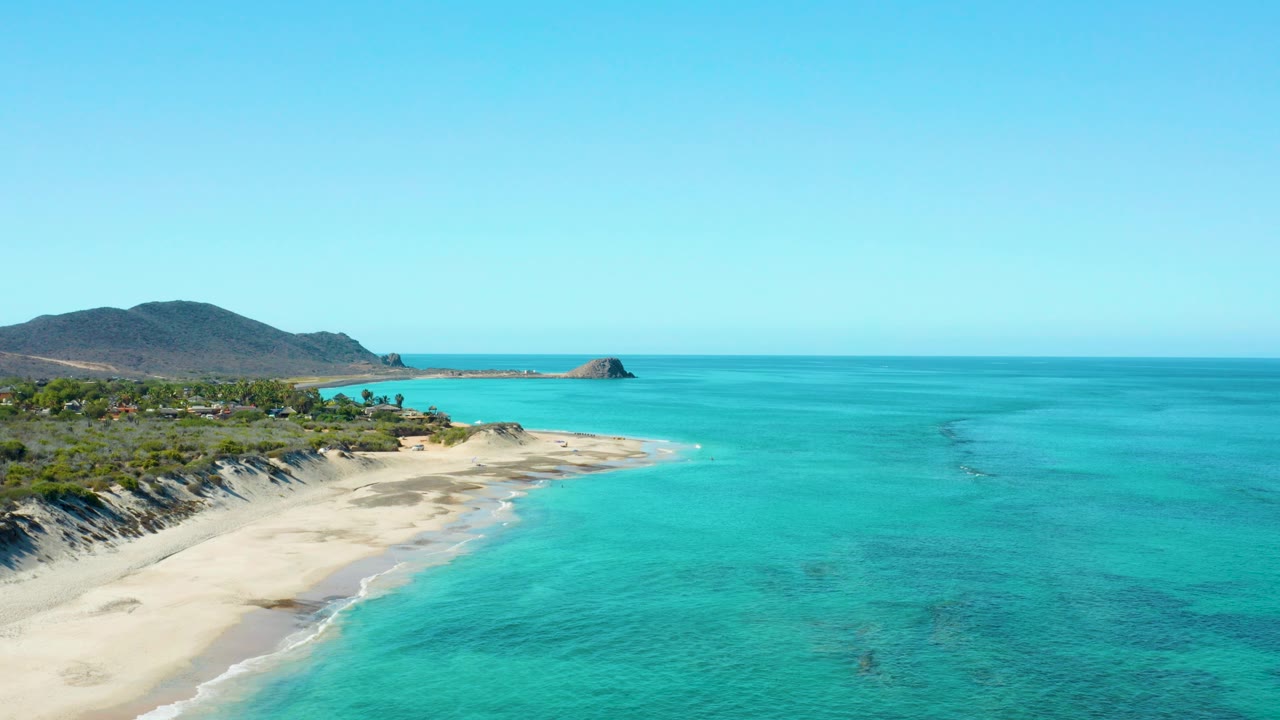 Opening aerial frame over Cabo Pulmo showing a curved sandy beach, bright turquoise shallows, green hillside, rocky point, clear blue sky, and no visible people.