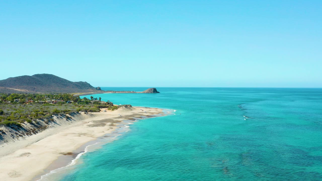 Cabo Pulmo coastline viewed from above with more open water on the right, the sandy beach at lower left, hills across the background, and no visible people.