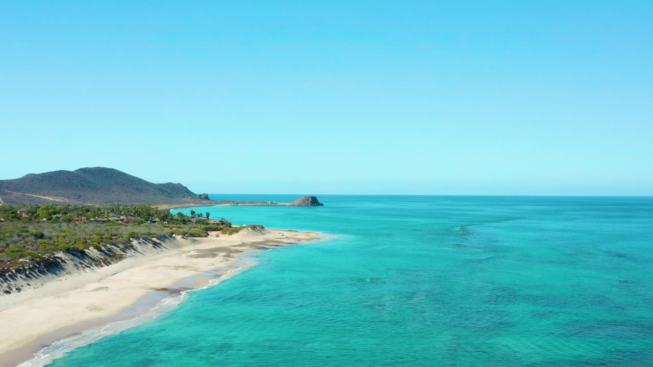 At 0:03, the frame shows the Cabo Pulmo shoreline curving through the left side with gentle waves, turquoise water stretching across the right side, hills in back, and no visible people.