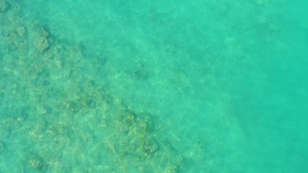 Clear Cabo Pulmo water seen from above, with a broad submerged rock patch entering from the upper left and faint ripples crossing the frame, no visible people.