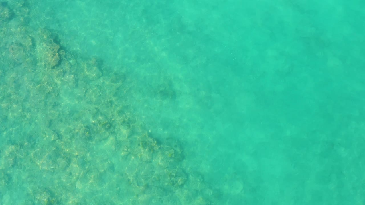 Overhead view of turquoise water in Cabo Pulmo, with rock and coral-like forms clustered near the upper left and center while the right side stays more open, no visible people.