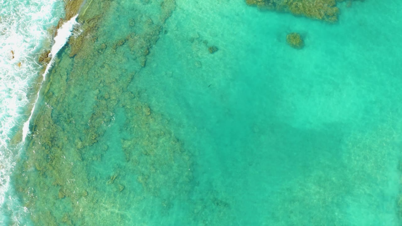 Clear turquoise shallows in Cabo Pulmo fill the frame, with foamy surf along the left edge and darker reef patches visible beneath the water, no visible people.