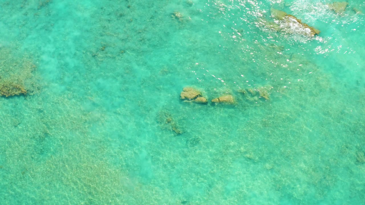 Overhead view of clear turquoise water in Cabo Pulmo with submerged rocks near the upper and middle frame, rippled sunlight, and no visible people.