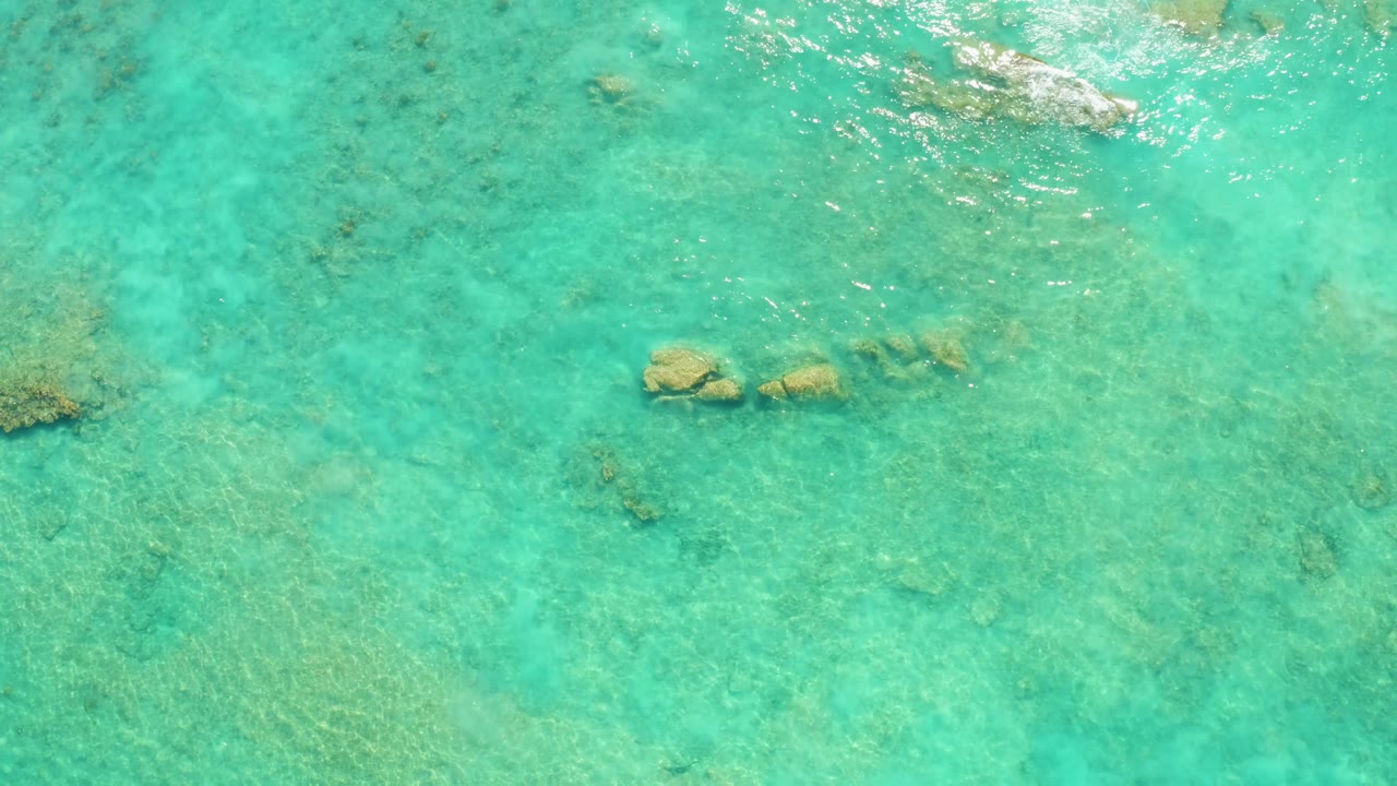 Overhead Cabo Pulmo frame with blue-green water, a brighter patch of sun reflection near the upper right rocks, scattered coral, and no visible people.