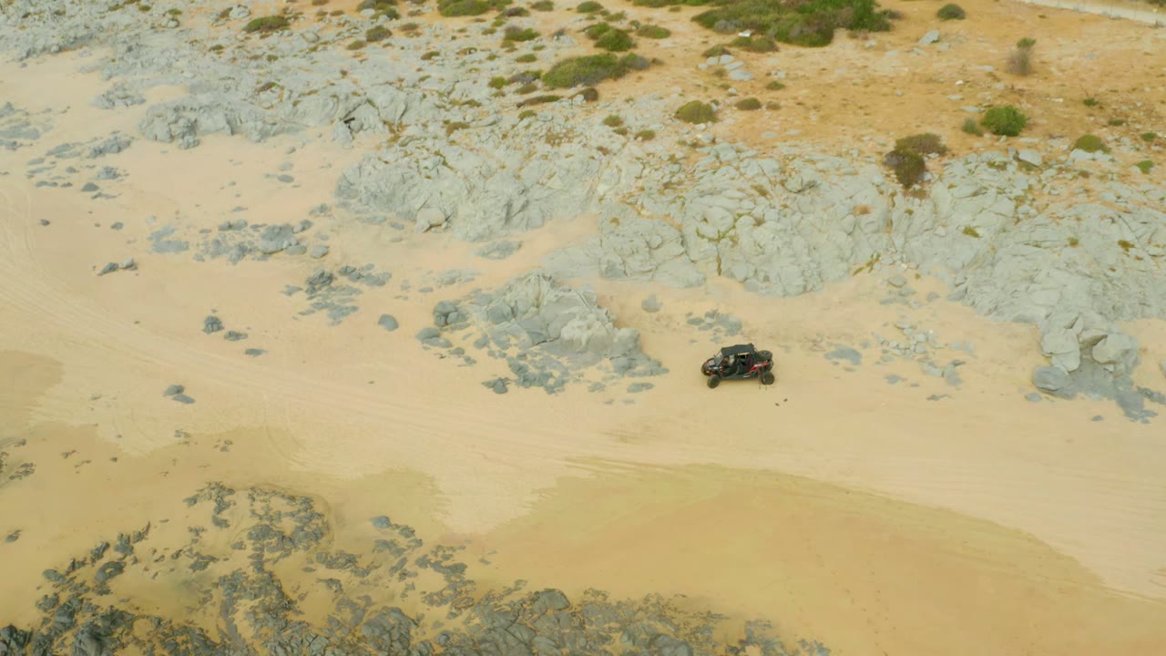 Overhead frame from Laguna Beach Los Cabos showing a dark 4x4 on pale sand beside gray rock outcrops, with no visible people.