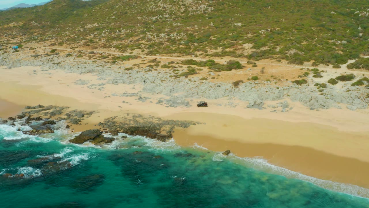 Aerial frame from Laguna Beach Los Cabos showing a dark 4x4 near the upper sand, turquoise surf and dark rocks in the foreground, scrub-covered hillside behind, and no visible people.