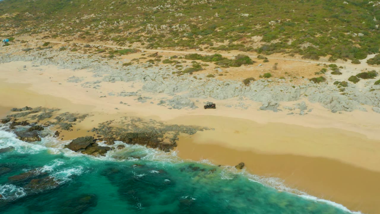Dark 4x4 on pale sand with rocky shoreline and clear blue-green water below.
