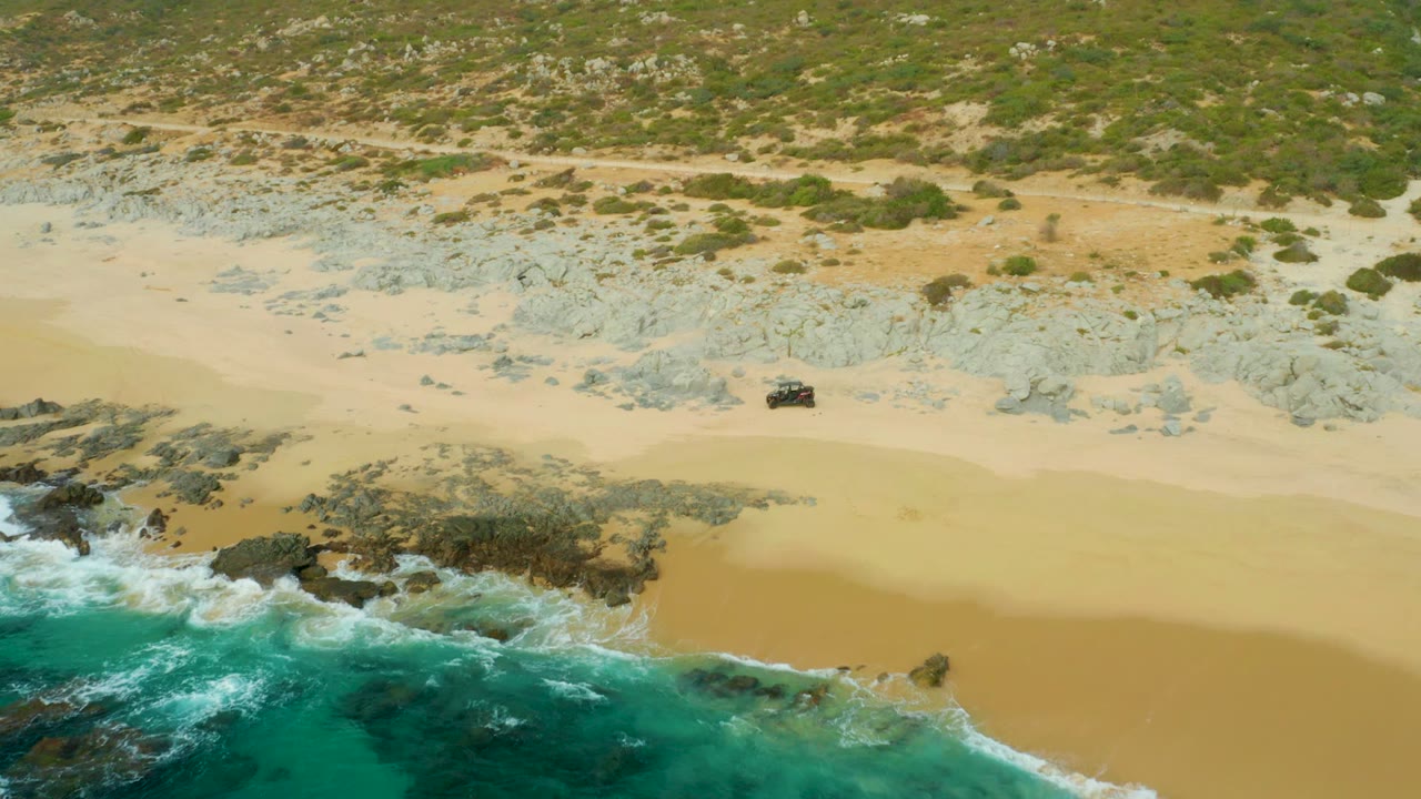Laguna Beach Los Cabos shoreline seen from above as the 4x4 continues across open sand, with foamy waves at the lower left, rock shelves near the water, and no visible people.