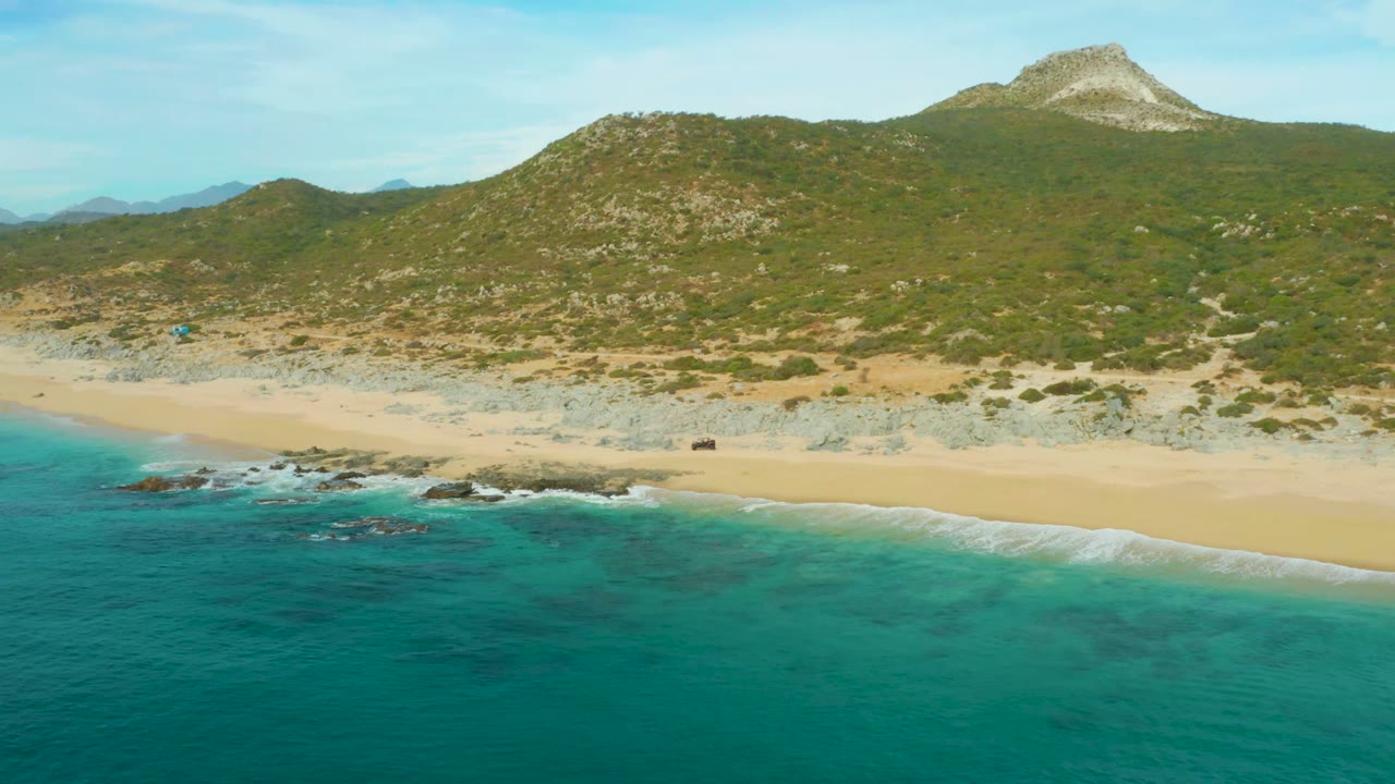 Aerial frame over Laguna Beach Los Cabos with turquoise ocean in front, pale sand and dark shoreline rocks in the middle, green hills and a distant peak behind, and no visible people.