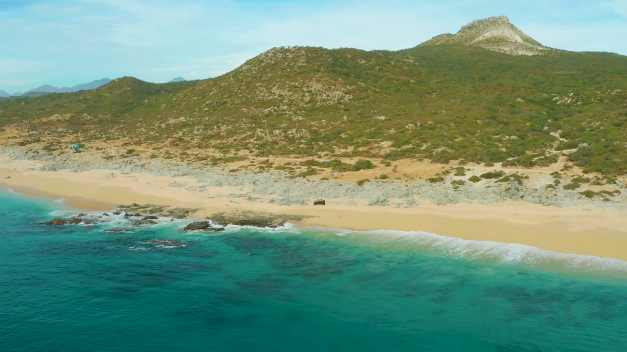 Turquoise ocean and surf below a sandy beach backed by rocky green hills.
