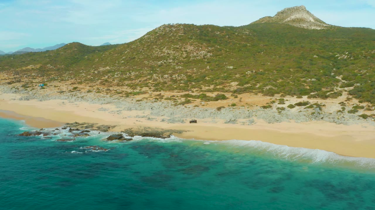 Wide aerial frame at Laguna Beach Los Cabos showing deeper turquoise water in the foreground, foam around shore rocks, a clean sand strip, green hillside, and no visible people.